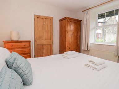 A bedroom with a bed, blue pillows, wooden wardrobe, chest of drawers, and window at Fisher's Retreat in Bowness-On-Windermere