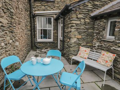 An outdoor stone patio with a blue table and chairs a white bench with two patterned cushions and a teapot with cups at Fishers Retreat in Bowness-On-Windermere