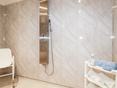 A shower area with a stainless steel shower panel a white chair and a white shelf with towels and a bottle at Chapel Green Studio 2 in Polgooth