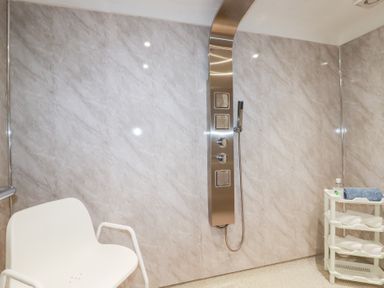 A bathroom with a white chair a stainless steel shower panel and a white plastic shelving unit with a blue towel and a small bottle at Chapel Green Studio 2 Polgooth