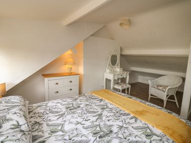 A bedroom with a bed with floral bedding a white dresser with lamp a white dressing table with stool and a wicker chair at Rowan Cottage in Builth Wells