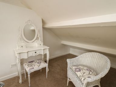 A small room with a white ornate dressing table with mirror a stool with floral upholstery and a white wicker chair with a floral cushion at Rowan Cottage in Builth Wells
