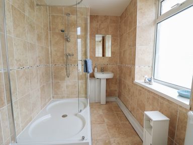 A bathroom with a corner shower, a pedestal sink, a mirror, a window sill with decorative items, and a small white cabinet at Rowan Cottage in Builth Wells