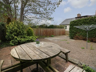 A backyard with a round wooden picnic table with a small watering can planter on top surrounded by gravel and greenery at Rowan Cottage in Builth Wells