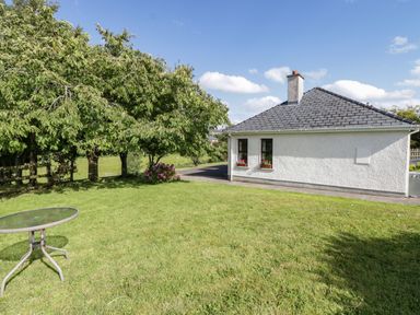 A garden with green grass a round glass table and a white house with flowers on windows at Belladrihid Cottage in Ballysadare near Sligo County Sligo