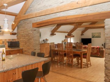 An open plan room with a kitchen island, dining table with chairs, sofas and exposed wooden beams at Cowslip Barn in Cornworthy near Dittisham