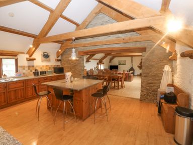 A kitchen with a granite island and bar stools next to a dining area with wooden chairs and exposed wooden beams at Cowslip Barn Cornworthy near Dittisham