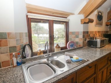 A kitchen sink with cleaning supplies and utensils on countertop and a window showing garden at Cowslip Barn in Cornworthy near Dittisham