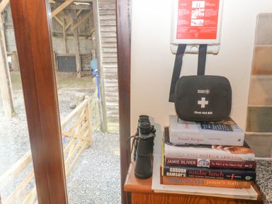 A corner with a stack of cookbooks a first aid kit and binoculars near a glass door at Cowslip Barn in Cornworthy near Dittisham