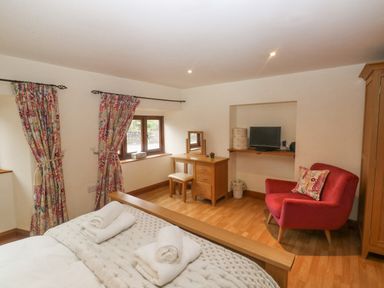 A bedroom with a bed, a wooden dressing table and chair, a red armchair, and colorful curtains at Cowslip Barn in Cornworthy near Dittisham