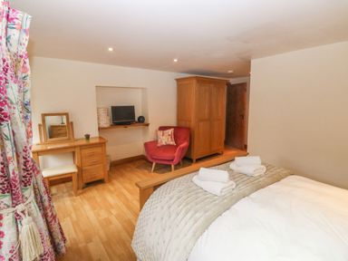 A bedroom with a bed covered with towels a wooden wardrobe a red armchair a dressing table and a TV on a wall shelf at Cowslip Barn in Cornworthy near Dittisham