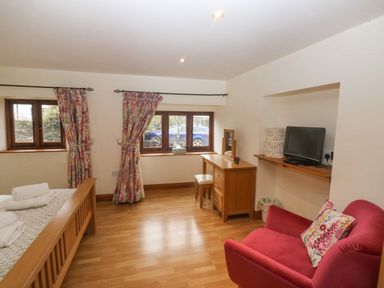 A bedroom with a wooden bed towels floral curtains desk chair red armchair and wall-mounted television at Cowslip Barn in Cornworthy near Dittisham