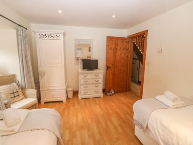A bedroom with two beds a white wardrobe a white chest of drawers with a TV a wicker chair and a wooden door at Cowslip Barn Cornworthy near Dittisham