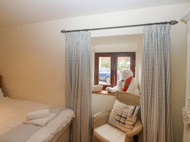 A bedroom with a bed and a chair next to a window with checkered curtains and a teddy bear on the window sill at Cowslip Barn in Cornworthy near Dittisham