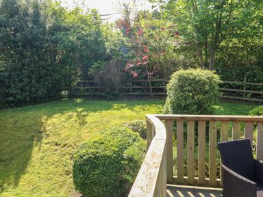 A garden with trimmed bushes trees and a wooden deck with a wicker chair at Cowslip Barn in Cornworthy near Dittisham