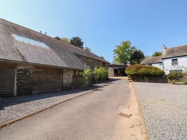 A driveway between stone and wooden barns with small trees and gravel at Cowslip Barn in Cornworthy near Dittisham