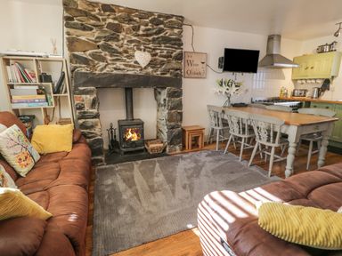 A living room with a stone fireplace a wood-burning stove brown sofas a dining table with chairs and a kitchen area at Snowdon View in Llanberis