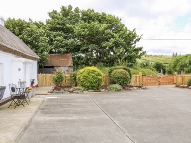 A concrete driveway with outdoor metal table and chairs next to a thatched white building and wooden gates in a rural landscape at Whispering Willows - The Thatch in Carndonagh County Donegal