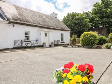 A white cottage with a thatched roof a small table and two chairs outside and flowers in pots in the foreground at Whispering Willows - The Thatch in Carndonagh County Donegal