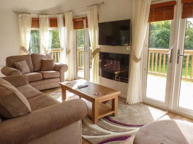 A living room with brown sofas a wooden coffee table a wall mounted TV and large windows with curtains at Morgan Lodge in Hewish near Weston-super-Mare