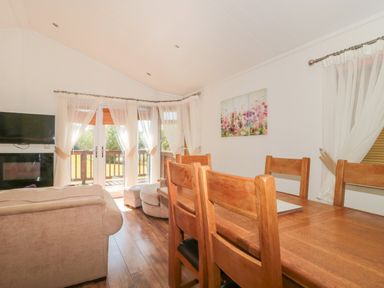 A dining area with a wooden table and chairs next to a living room with a beige sofa and a window with sheer curtains at Harvester Lodge in Hewish near Weston-super-Mare