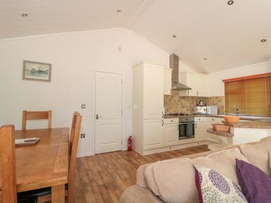 An open plan kitchen and dining area with wooden dining table and chairs beige kitchen cabinets and a window with blinds at Harvester Lodge in Hewish near Weston-super-Mare