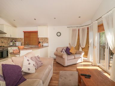 A living room with two beige sofas and a wooden coffee table next to a kitchen with tiled backsplash and wooden blinds at Harvester Lodge in Hewish near Weston-super-Mare