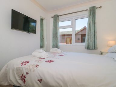 A bedroom with a bed towels a wall-mounted television window with curtains and a lamp at Harvester Lodge in Hewish near Weston-super-Mare