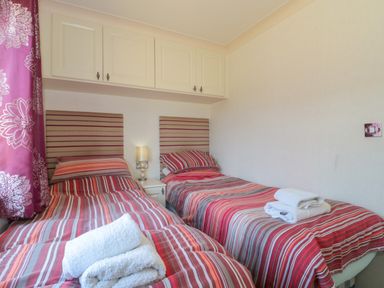 A bedroom with two single beds striped bedding striped headboards a bedside table with a lamp and overhead cupboards at Harvester Lodge in Hewish near Weston-super-Mare