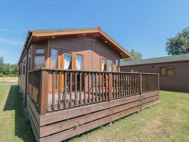 A wooden lodge with a fenced deck and multiple windows on a grassy area at Harvester Lodge in Hewish near Weston-super-Mare
