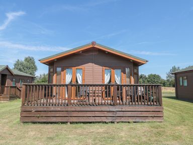A wooden cabin with a front deck and railing on a grassy area under a blue sky at Harvester Lodge in Hewish near Weston-super-Mare