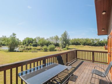 A wooden deck with a table and chairs overlooking a grassy area with trees and picnic benches at Harvester Lodge in Hewish near Weston-super-Mare