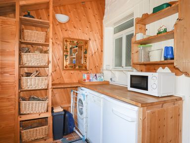 A utility room with wooden shelves holding baskets and a microwave on a wooden counter at The Ark Cottage in St Blazey