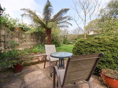 A garden patio with two chairs and a round glass table surrounded by plants and trees at The Ark Cottage in St Blazey