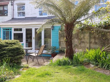 A garden with a tree a bush and a chair and table set outside a house with glass doors at The Ark Cottage in St Blazey