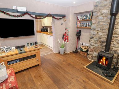 A living room with a wood stove fire a wooden cabinet with framed photos a television and an open doorway to a kitchen at Stable Cottage in Pembroke