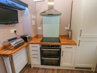A kitchen area with an electric stove and oven a toaster a bread box a TV mounted on the wall and cutting boards at Stable Cottage in Pembroke