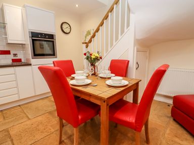 A dining area with a wooden table set for four with red chairs and a kitchen with white cabinets and an oven at Cow Byre Great Tew near Chipping Norton