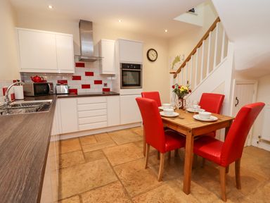 A kitchen with white cabinets a wooden dining table with red chairs and a staircase at Cow Byre Great Tew near Chipping Norton