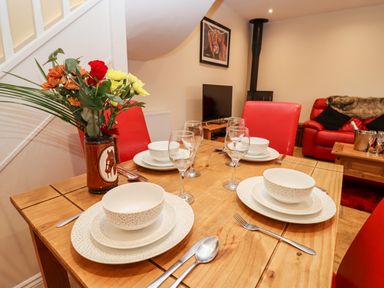 A dining table set with plates bowls cutlery and glasses with a vase of flowers in a room with red chairs a red sofa a TV and a wood burner at Cow Byre Great Tew near Chipping Norton