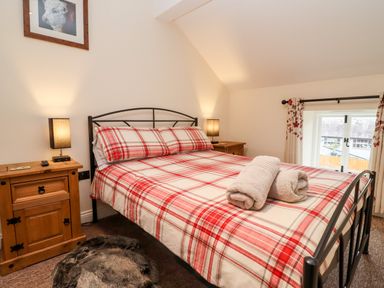 A bedroom with a double bed with red plaid bedding two wooden bedside tables with lamps and a small window with floral curtains at Cow Byre Great Tew near Chipping Norton