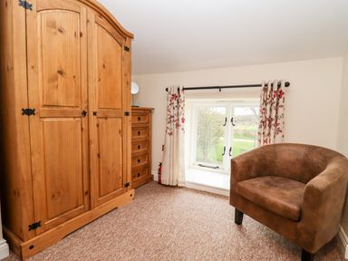 A room with a wooden wardrobe chest of drawers and a brown armchair next to a window with curtains at Cow Byre Great Tew near Chipping Norton