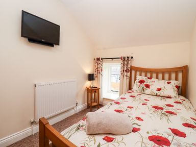 A small bedroom with a wooden bed with floral bedding a bedside table with a lamp a wall-mounted TV and a small window at Cow Byre Great Tew near Chipping Norton