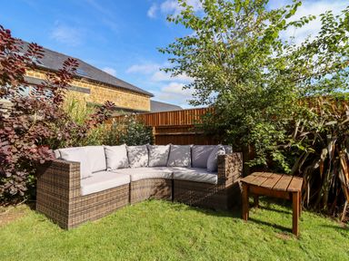 An outdoor corner sofa with cushions and a wooden table on grass surrounded by plants at Cow Byre Great Tew near Chipping Norton
