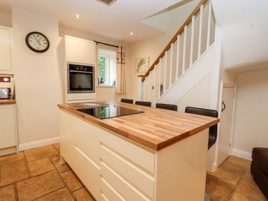 A kitchen with a wooden countertop island black chairs under stairs and a wall clock at The Stables in Great Tew near Chipping Norton