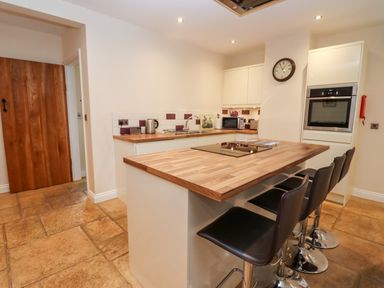 A kitchen with a wooden island counter with three black bar stools a built in oven microwave wall clock and wooden door at The Stables in Great Tew near Chipping Norton