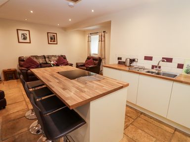 A kitchen area with a wooden countertop island and bar stools next to a living space with leather sofas at The Stables in Great Tew near Chipping Norton