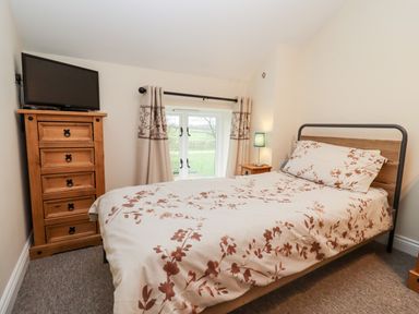 A bedroom with a bed covered in floral bedding a wooden chest of drawers with a television on top and a window with curtains at The Stables in Great Tew near Chipping Norton