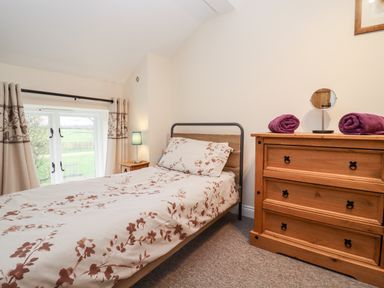 A bedroom with a single bed covered with floral bedding a wooden dresser with two purple towels and a mirror and a window with curtains at The Stables Great Tew near Chipping Norton