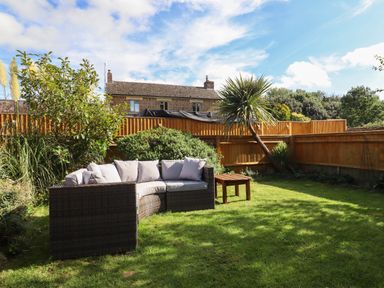 A garden with a wicker sofa and cushions a wooden table a tree a fence and a house in the background at The Stables in Great Tew near Chipping Norton